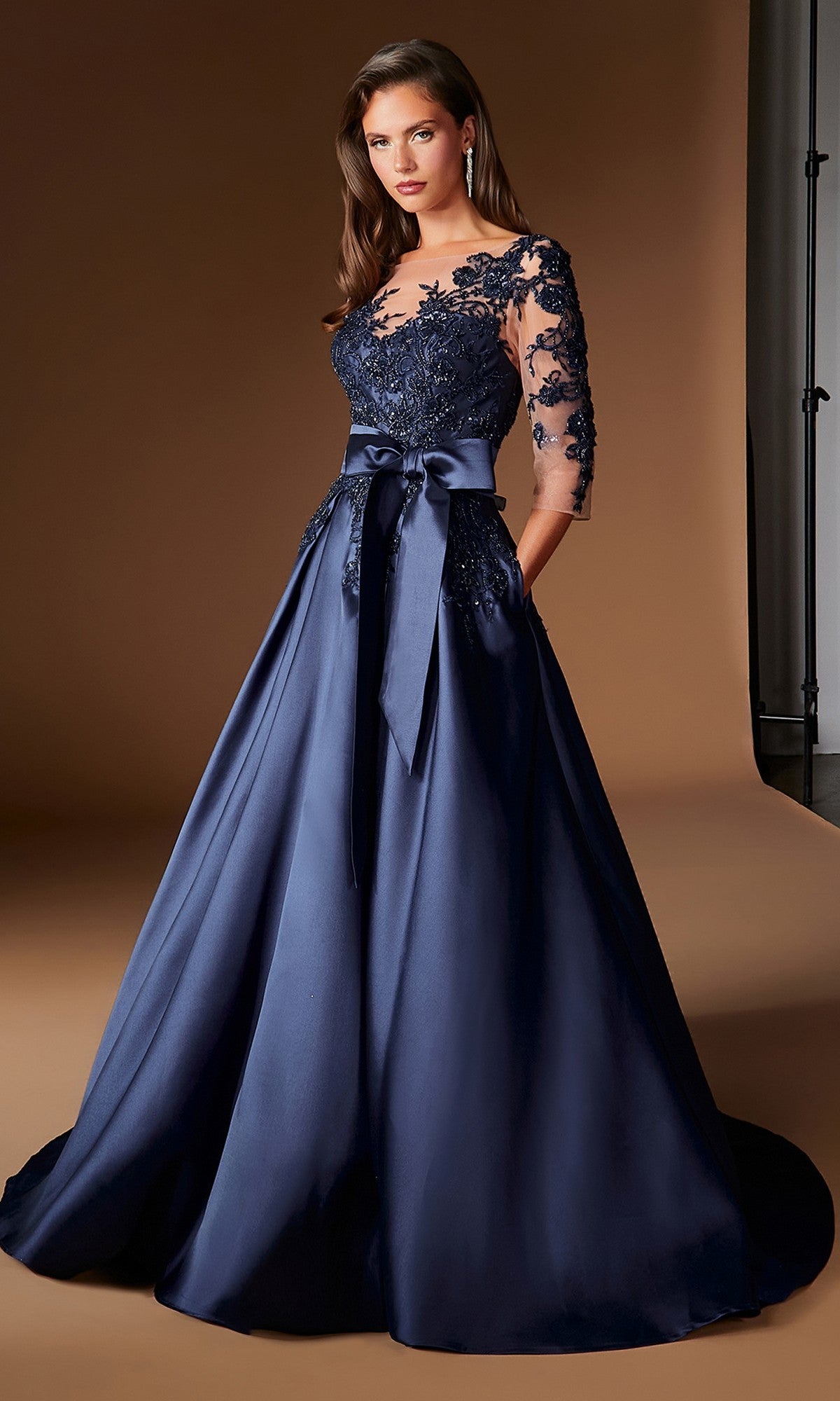 A woman stands in a studio setting wearing the Ladivine CDS569 long formal dress in navy, featuring embroidered lace, an appliqué bodice, sheer sleeves, and a floor-length silhouette finished with a satin sash.